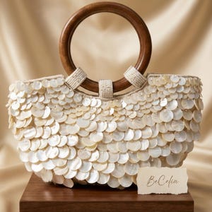 Op de afbeelding: Een witte handtas bedekt met schelpen met een houten handvat. De tas heeft een cirkelvormig houten handvat en is versierd met talrijke kleine, ronde, witte schelpen. De merknaam "BeCelia" staat op een klein kaartje.