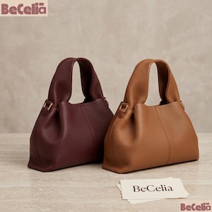 May include: Two leather handbags in burgundy and tan. Each bag has a rounded shape with a top handle and gold-colored hardware. The bags are displayed on a light-colored surface. The brand name "BeCelia" is visible in the upper left and lower right corners, and on a small card.