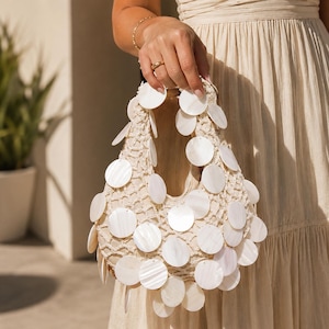 May include: A cream-colored crochet handbag featuring a structured net design adorned with numerous circular, iridescent white shell discs attached across the exterior surface, creating a layered and textured appearance on the small tote bag.
