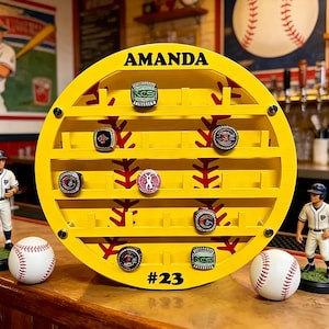 May include: A yellow baseball ring display with the name "AMANDA" at the top and the number "23" at the bottom. The display holds several rings and is decorated with red baseball stitching. Two baseball figurines and two baseballs are also present.