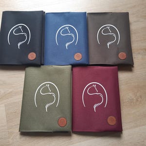 Könnte beinhalten: Fünf Stoffnotizbücher in Schwarz, Marineblau, Braun, Olivgrün und Burgunderrot. Jedes Notizbuch hat ein weißes Pferdekopfdesign in einem Kreis und einen kleinen, runden, braunen Lederaufnäher. Auf Holz angeordnet.