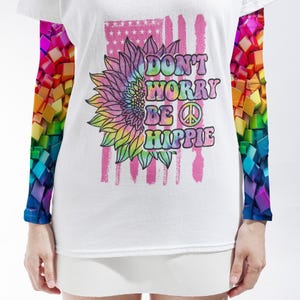 May include: A white long-sleeve t-shirt featuring a tie-dye sunflower and a peace sign graphic with the text DON'T WORRY BE HIPPIE. The sleeves display a vibrant, multi-colored 3D cube pattern, while the center shows a faded pink American flag.