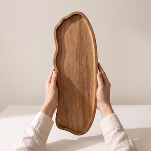 May include: An elongated wooden serving tray with a natural light brown wood grain and an irregular, organic wavy edge. The tray features a smooth, flat surface with a slightly raised rim, showcasing the distinct texture and patterns of the wood.