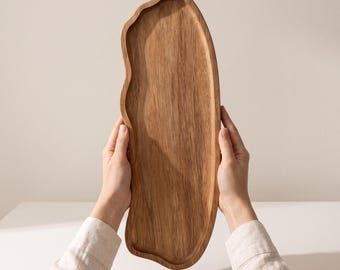 May include: An elongated wooden serving tray with a natural light brown wood grain and an irregular, organic wavy edge. The tray features a smooth, flat surface with a slightly raised rim, showcasing the distinct texture and patterns of the wood.