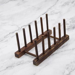 Può includere: Uno scolapiatti in legno marrone scuro caratterizzato da due barre di base rettangolari parallele collegate da un'asta di supporto centrale, con dieci pioli cilindrici verticali che si estendono verso l'alto per sostenere piatti.
