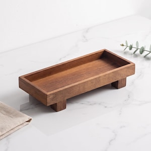 May include: A rectangular wooden tray with a smooth, dark brown finish and visible wood grain. The tray features raised edges and is supported by four short, block-style feet at each corner, elevating the base slightly above the surface.
