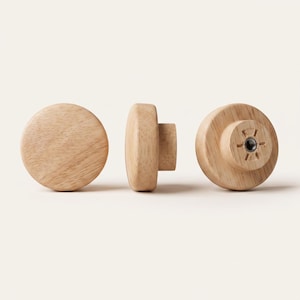 Puede incluir: Tres pomos de madera de color claro con una cara circular lisa y un patrón de veta natural. Cada pomo presenta una base cilíndrica con un inserto roscado de metal en el centro de la parte posterior, diseñado para fijarse a muebles o armarios.
