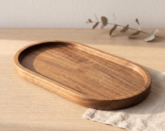 Könnte beinhalten: Ein ovales Holztablett mit einer glatten, polierten Oberfläche und sichtbaren natürlichen Holzmaserungen. Das Tablett verfügt über eine flache Vertiefung mit erhöhten Rändern und zeigt einen warmen, mittelbraunen Farbton über die gesamte Struktur.