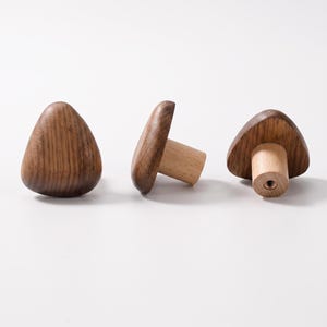 Puede incluir: Tres pomos de madera para armarios con una cabeza triangular de color marrón oscuro con vetas suaves y redondeadas, y una base cilíndrica de color claro. La base del pomo de la derecha muestra un orificio central de metal.