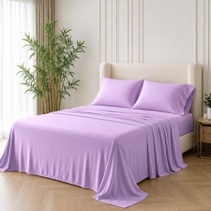 May include: A bed featuring a solid light purple flat sheet and two matching light purple pillowcases. The bedding is draped over a mattress on a light-coloured upholstered bed frame with a tall headboard, creating a smooth and uniform appearance.