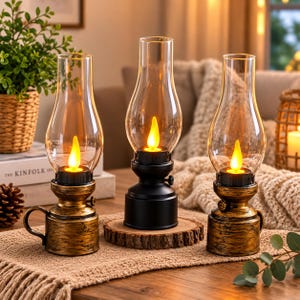 Può includere: Tre lampade in stile olio con camini in vetro trasparente e fiamme LED gialle luminose. Due lampade hanno una finitura oro anticato con manici laterali, mentre la lampada centrale ha una base nera opaca e poggia su un disco di legno.