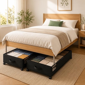 May include: A light oak wooden bed frame with a white duvet and a green accent pillow. Two black fabric under-bed storage drawers with wheels and handles are pulled out, each containing neatly folded clothing items in various colors.