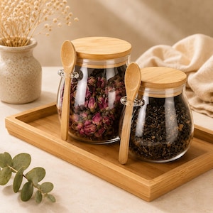 May include: Two clear glass jars with round bamboo lids, each featuring a small bamboo spoon held by a glass loop on the side. One jar is filled with dried pink rose buds, and the other contains dark loose-leaf tea, both resting on a bamboo tray.