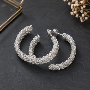 May include: A pair of silver hoop earrings featuring a textured, woven mesh design. The earrings have a circular shape with a metallic finish and include a post-back closure mechanism, showcasing a detailed, intricate pattern across the entire surface.