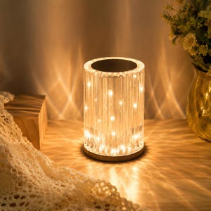 May include: A cylindrical table lamp with a clear, vertically ribbed acrylic body that emits a warm, glowing light. The lamp features a flat, dark circular base and top, casting intricate, diamond-shaped light patterns onto the surrounding area.
