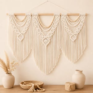 May include: A large off-white macrame wall hanging suspended from a wooden dowel, featuring three symmetrical sections with intricate knotted patterns, draped fringe, and integrated light-colored wooden beads in a curved arrangement.