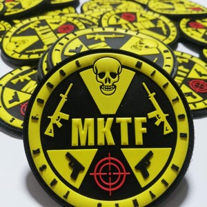 Puede incluir: Un conjunto de parches redondos, negros y amarillos. Cada parche presenta una calavera, armas y el texto "MKTF". Los parches tienen un símbolo de diana y el texto "JUST IN SINA".