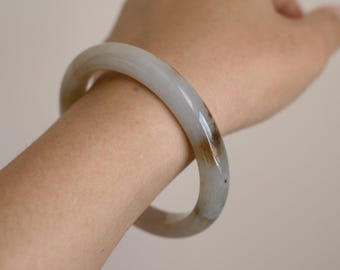 May include: A smooth, polished bangle bracelet made of translucent light gray stone with subtle, cloudy white inclusions and a distinct patch of warm brown coloration integrated into the circular band, worn around a wrist.