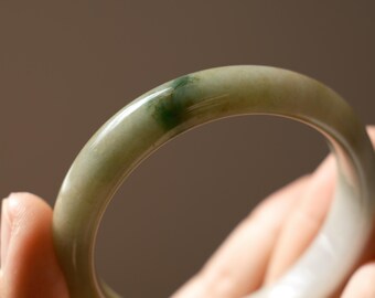 May include: A smooth, polished jade bangle bracelet featuring a translucent pale green base with a distinct, concentrated patch of deep emerald green color. The circular jewelry piece has a rounded profile and a glossy, reflective surface finish.