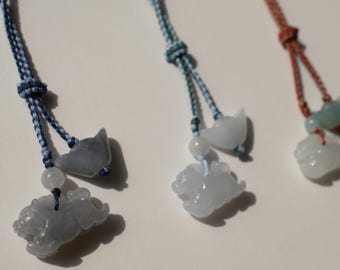 May include: Close-up of several jade pendants with animal carvings. Each pendant is suspended from a colorful braided cord. The pendants feature a variety of animal shapes, including a cat and a pig. The jade is a pale green color.