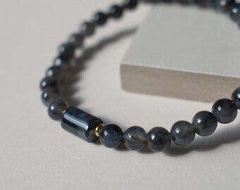 May include: A beaded bracelet featuring smooth, dark grey, spherical stones arranged in a circular pattern. The design includes a single, elongated, cylindrical dark grey bead accented by a small, polished gold-toned metal spacer ring.