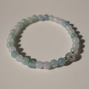 May include: A beaded bracelet featuring smooth, round, translucent stones in shades of pale green, soft blue, and milky white. The circular strand is accented by a single larger, slightly darker grey-toned bead flanked by two small silver-toned metal spacers.