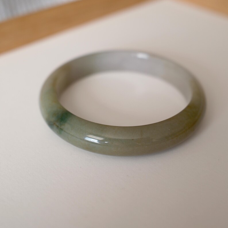 May include: A smooth, polished jade bangle bracelet featuring a rounded profile. The stone displays a translucent, pale greyish-green base color with a distinct, darker emerald green patch integrated into the natural mineral structure of the band.