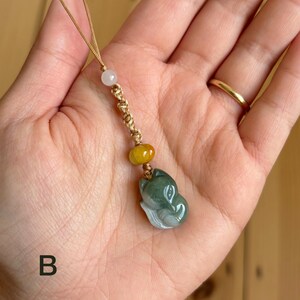 May include: A jade pendant featuring a carved fox head in shades of green and white. The pendant is strung on a gold-colored cord with a white bead and a yellow bead. The letter "B" is visible in the lower left corner.