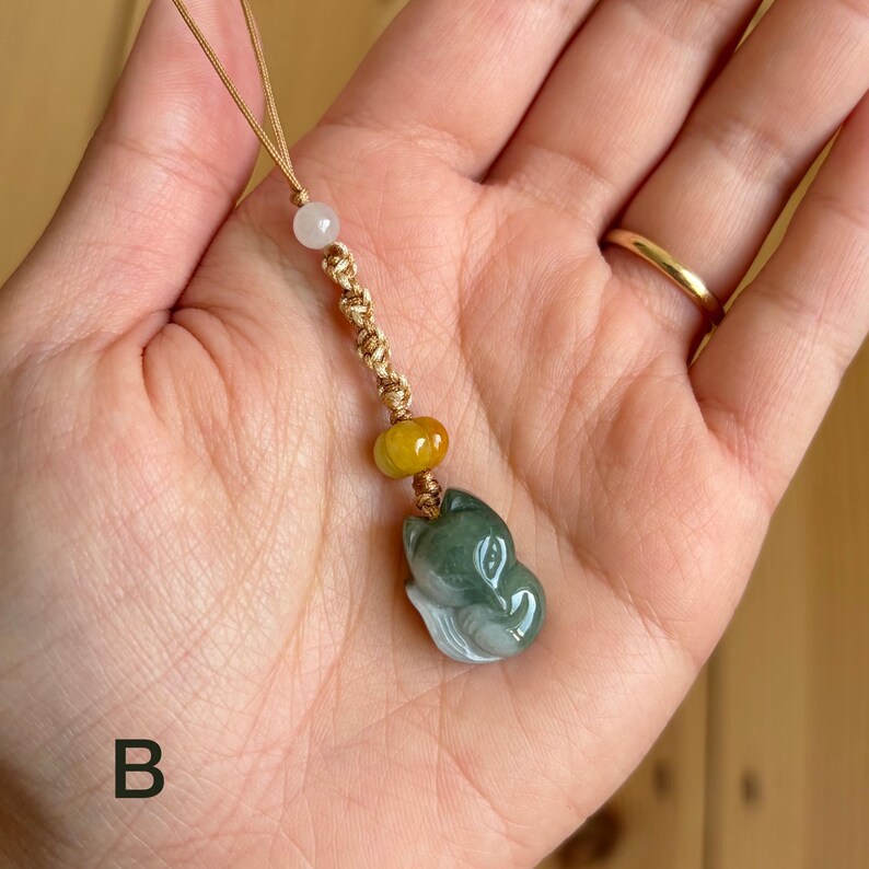 May include: A jade pendant featuring a carved fox head in shades of green and white. The pendant is strung on a gold-colored cord with a white bead and a yellow bead. The letter "B" is visible in the lower left corner.
