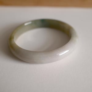 May include: A smooth, polished bangle bracelet crafted from stone, featuring a rounded profile with a translucent appearance. The surface displays a gradient of colors, transitioning from soft white and pale lavender to patches of muted green.