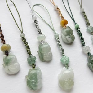 May include: A collection of jade fox pendants with various cord colors. Each pendant features a carved fox figure in shades of green and white, accented with beads and decorative knots. The pendants are designed for use as charms or accessories.