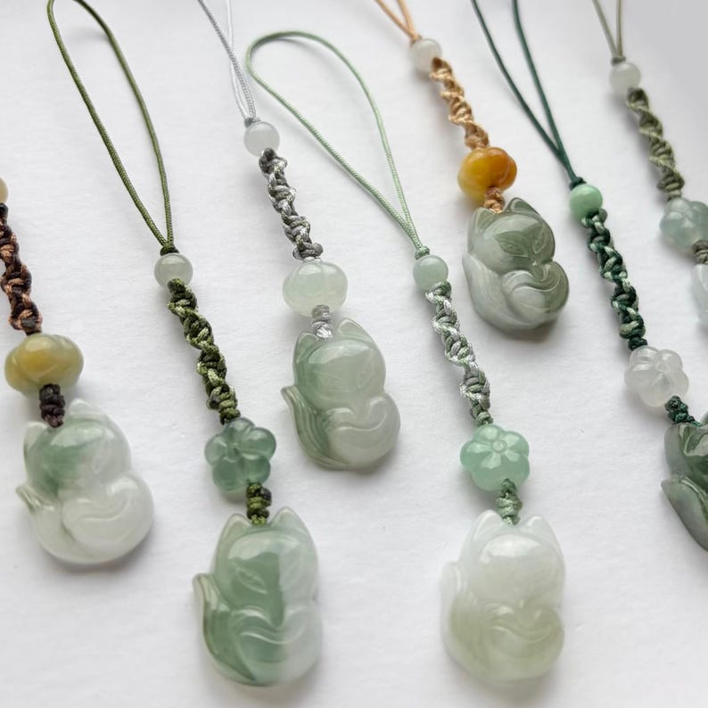 May include: A collection of jade fox pendants with various cord colors. Each pendant features a carved fox figure in shades of green and white, accented with beads and decorative knots. The pendants are designed for use as charms or accessories.