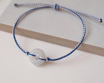 May include: A bracelet featuring a smooth, pale green circular stone bead centered on a thin, braided cord. The cord is constructed from intertwined white and royal blue threads, with adjustable ends extending from a small central knot.