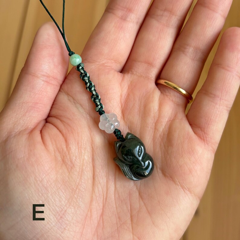 May include: A dark green jade fox pendant with a black and green braided cord, a light green bead, and a clear flower-shaped bead. The fox pendant is intricately carved and has a glossy finish. The cord is attached to a black string.