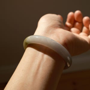 May include: A smooth, polished bangle bracelet crafted from translucent, light gray stone with a subtle hint of olive green near one edge. The circular accessory features a rounded profile and a glossy, reflective surface that catches the light.