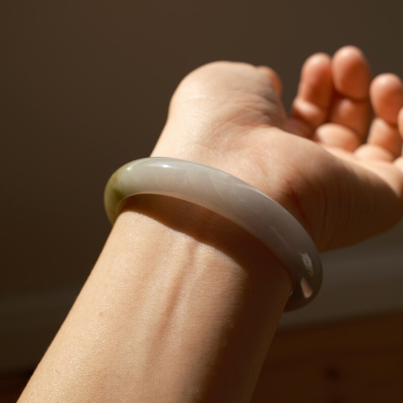 May include: A smooth, polished bangle bracelet crafted from translucent, light gray stone with a subtle hint of olive green near one edge. The circular accessory features a rounded profile and a glossy, reflective surface that catches the light.
