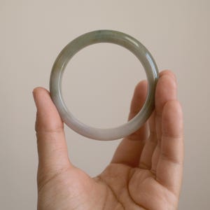 May include: A smooth, circular bangle bracelet crafted from polished stone. The bangle features a translucent appearance with a gradient of soft sage green and muted grey tones, showcasing natural variations in the stone's color and texture.
