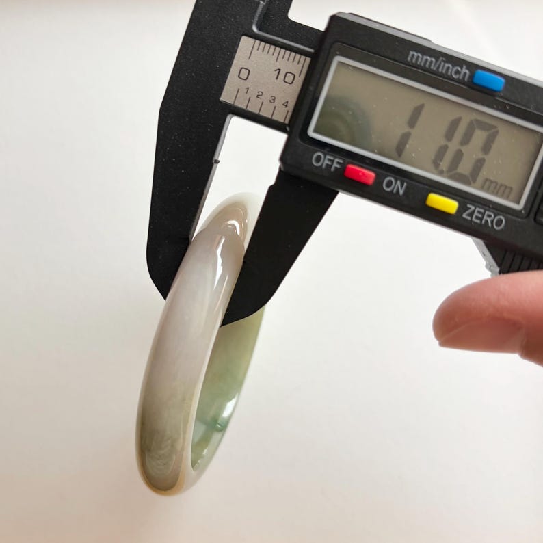 May include: A smooth, pale green and white translucent bangle bracelet being measured by a black digital caliper. The digital display on the caliper clearly shows a measurement of 11.0 mm, indicating the thickness of the stone band.