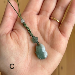 May include: A jade pendant with a carved fox design, suspended from a green cord. The pendant is a pale green color with darker green accents. A small flower and bead accent the cord.