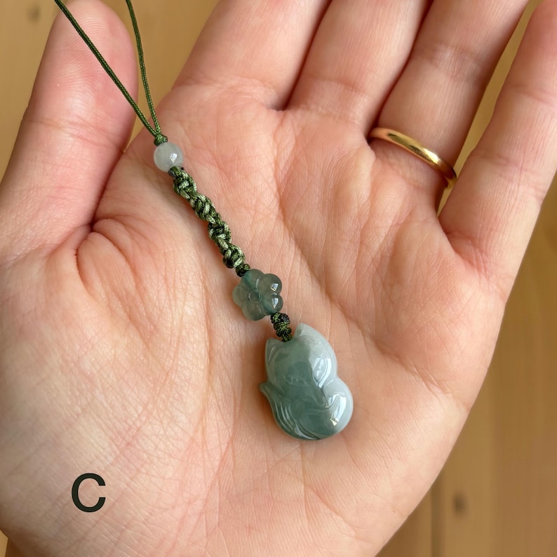 May include: A jade pendant with a carved fox design, suspended from a green cord. The pendant is a pale green color with darker green accents. A small flower and bead accent the cord.