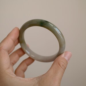 May include: A smooth, polished bangle bracelet crafted from translucent stone with a rounded profile. The material displays a soft, muted color palette featuring blended shades of pale grey, beige, and a distinct deep green patch.