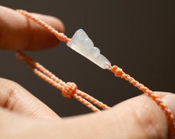 May include: A bracelet featuring a pale, translucent, white stone charm with a stepped, tiered design. The charm is attached to a braided, peach-colored cord that includes adjustable sliding knots for sizing, creating a simple and functional accessory.