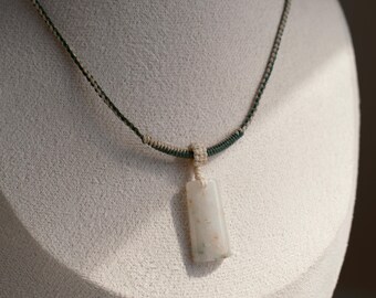 May include: A rectangular pendant necklace with a cream-colored stone. The stone is suspended from a macrame cord with a green and beige pattern. The necklace is displayed on a white mannequin head.