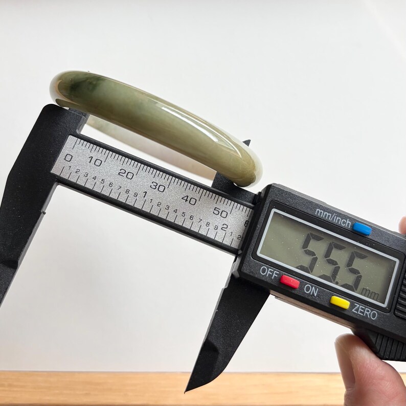 May include: A smooth, polished, translucent green jade bangle bracelet being measured by a black digital caliper. The digital display clearly shows a measurement of 55.5 mm, indicating the inner diameter of the circular stone jewelry piece.