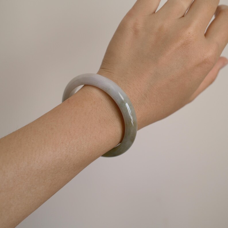 May include: A smooth, solid bangle bracelet crafted from polished stone. The accessory features a rounded, continuous circular band with a translucent, pale greyish-green hue and subtle variations in color throughout the natural material.