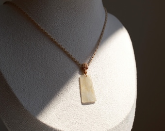 May include: A beige rectangular pendant necklace. The pendant is a light, creamy color and hangs from a beige cord. The necklace is displayed on a white mannequin. The background is a soft, neutral tone.
