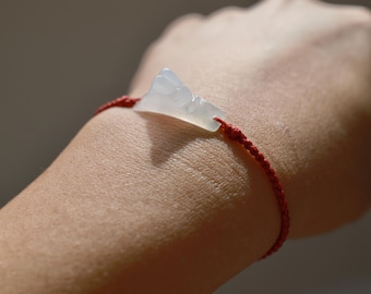 May include: A bracelet featuring a translucent, pale white carved stone charm shaped like a small animal, secured by a thin, braided red cord. The charm is centered on the cord, which wraps around the wrist with adjustable knots on each side.