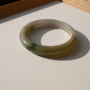 May include: A smooth, circular bangle bracelet crafted from translucent stone with a polished finish. The piece features a gradient of muted green and pale grey tones, with a distinct darker green inclusion visible along one side of the band.