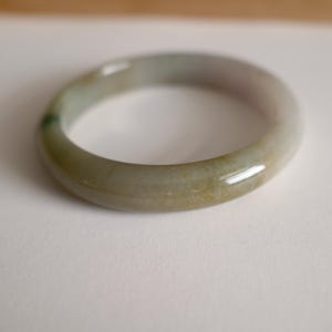 May include: A smooth, polished bangle bracelet crafted from translucent stone with a rounded profile. The material displays a variegated appearance, featuring soft blends of pale green, muted grey, and subtle earthy beige tones throughout the surface.