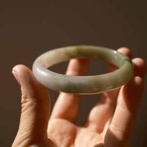 May include: A smooth, circular bangle bracelet crafted from translucent stone with a polished finish. The material displays a variegated appearance, featuring soft gradients of pale grey, off-white, and subtle patches of muted forest green.